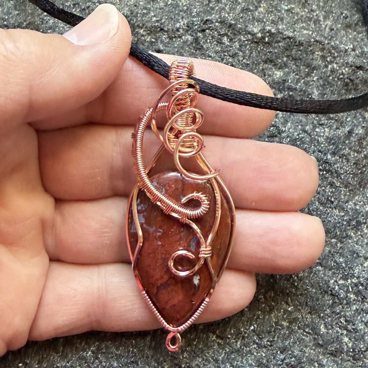 Front view of an inverted-teardrop shaped red plume agate pendant wrapped in copper wire and suspended from a black cord, shown held in the artist's hand.
