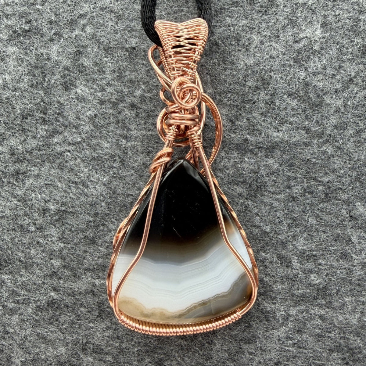 Back view of a teardrop shaped Botswana agate wrapped in copper wire, held by a black cord on a gray felt backdrop.