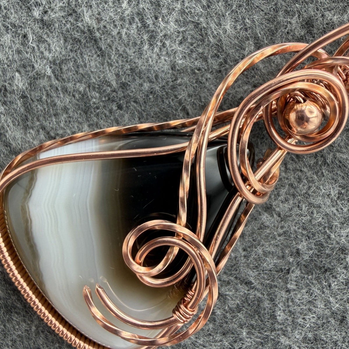 Detail view of the wire-wrapping on a teardrop shaped Botswana agate wrapped in copper wire, held by a black cord on a gray felt backdrop.