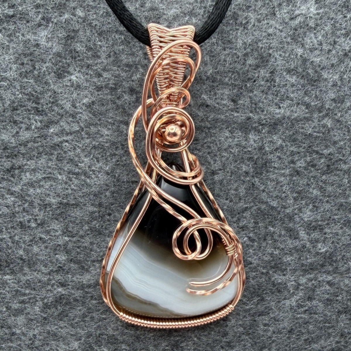 Front view of a teardrop shaped Botswana agate wrapped in copper wire, held by a black cord on a gray felt backdrop.