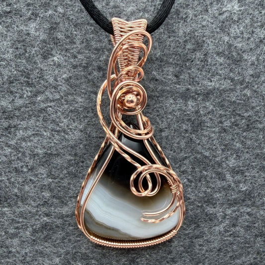 Front view of a teardrop shaped Botswana agate wrapped in copper wire, held by a black cord on a gray felt backdrop.