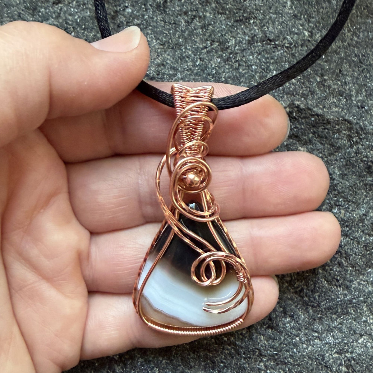 Front view of a teardrop shaped Botswana agate wrapped in copper wire, held by a black cord, resting in the artist's hand.