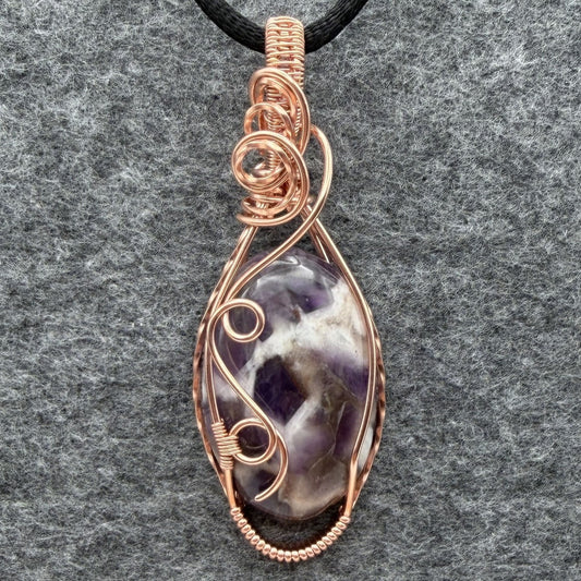 Front view of an oval dream amethyst pendant wrapped in copper wire, hanging from a black satin cord against a gray felt background.