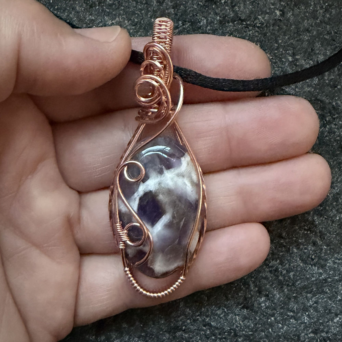 Front view of an oval dream amethyst pendant wrapped in copper wire, hanging from a black satin cord held in the artist's hand.