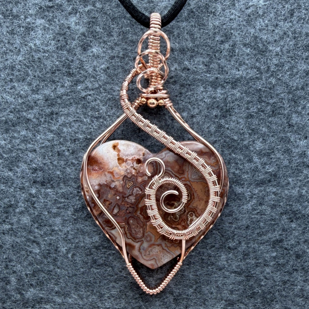 Front view of a pink crazy lace agate heart wrapped in copper wire suspended from a black cord against a gray felt background.