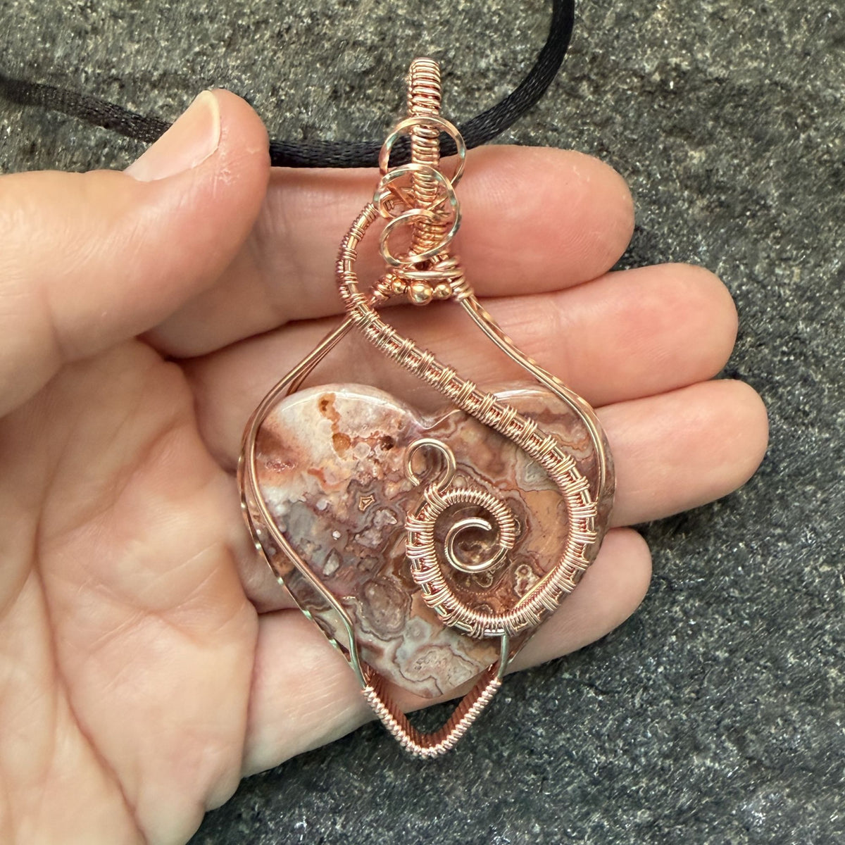 Front view of a pink crazy lace agate heart wrapped in copper wire suspended from a black cord held in the artist's hand.