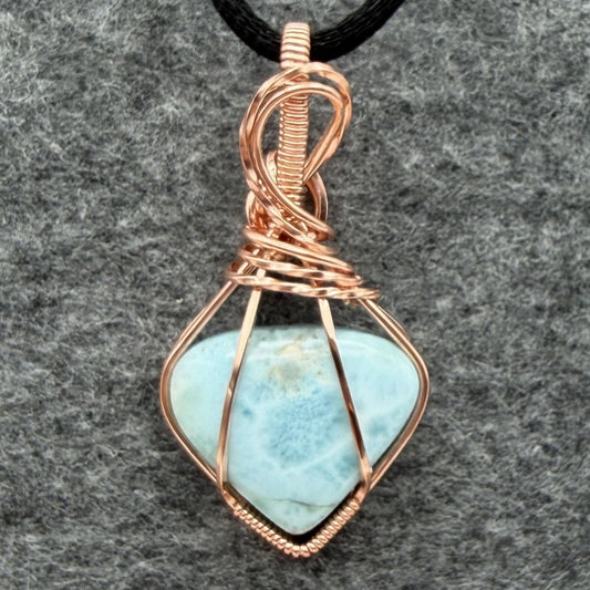 Front view of a pale blue larimar pendant wrapped in polished copper wire with a looping bail, hanging from a black cord against a gray felt background.