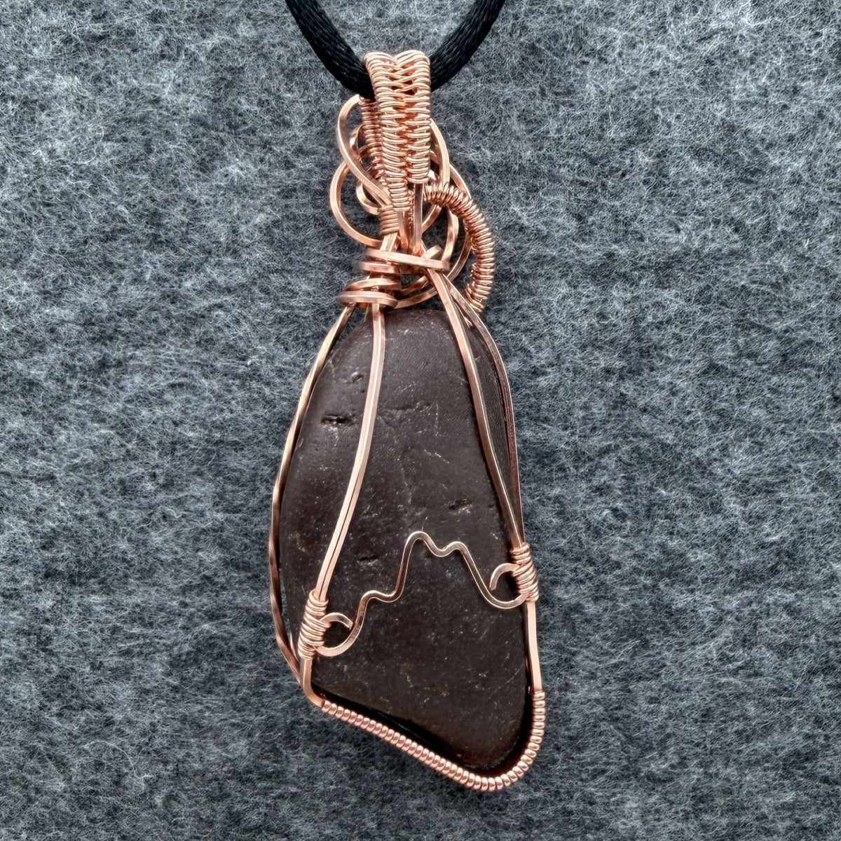 Back view of a natural stone pendant wrapped in copper wire, hanging from a black cord, against a gray felt background.