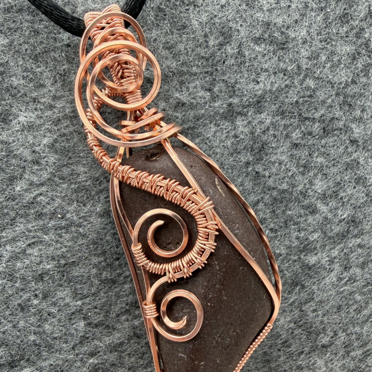 Detail view of the front of a natural stone pendant wrapped in copper wire, hanging from a black cord, against a gray felt background.