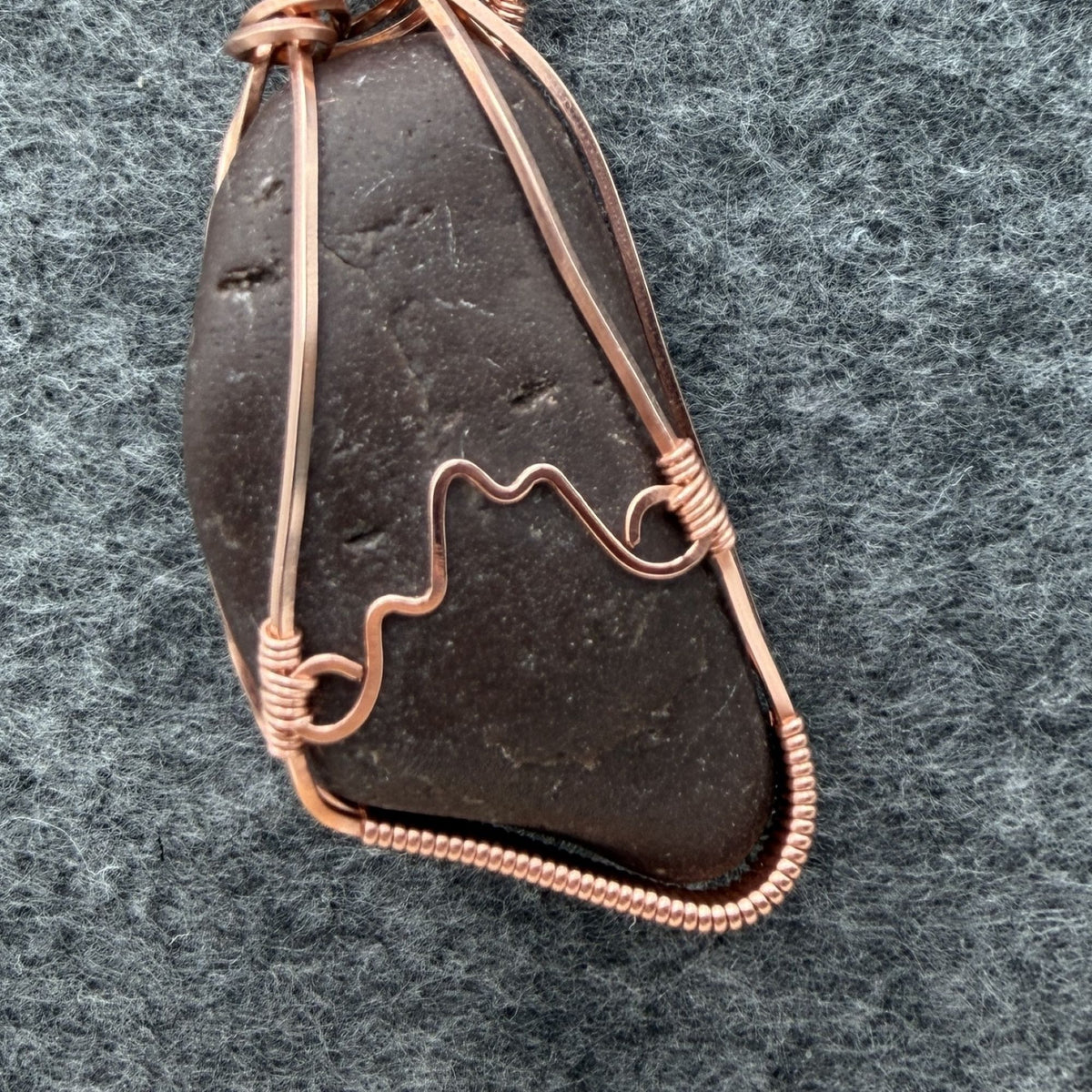 Detail view of the back of a natural stone pendant wrapped in copper wire, hanging from a black cord, against a gray felt background.