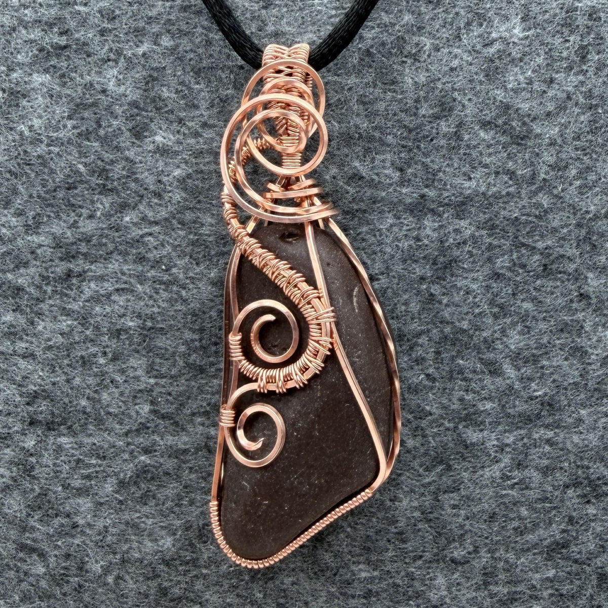 Front view of a natural stone pendant wrapped in copper wire, hanging from a black cord, against a gray felt background.