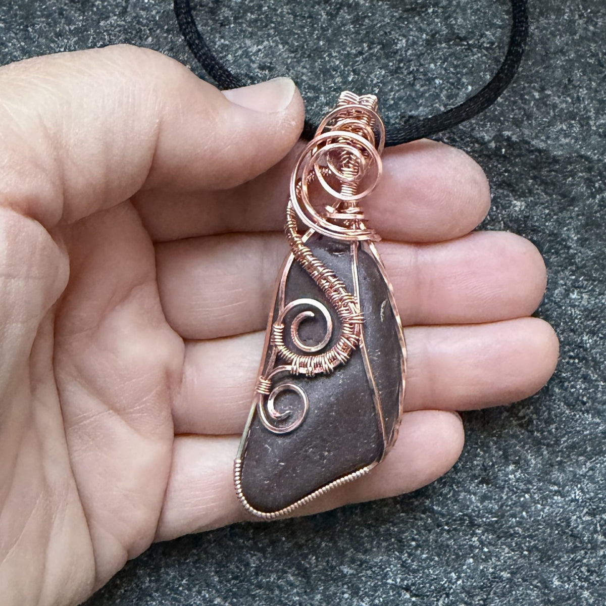 Front view of a natural stone pendant wrapped in copper wire, hanging from a black cord, held in the artist's hand.
