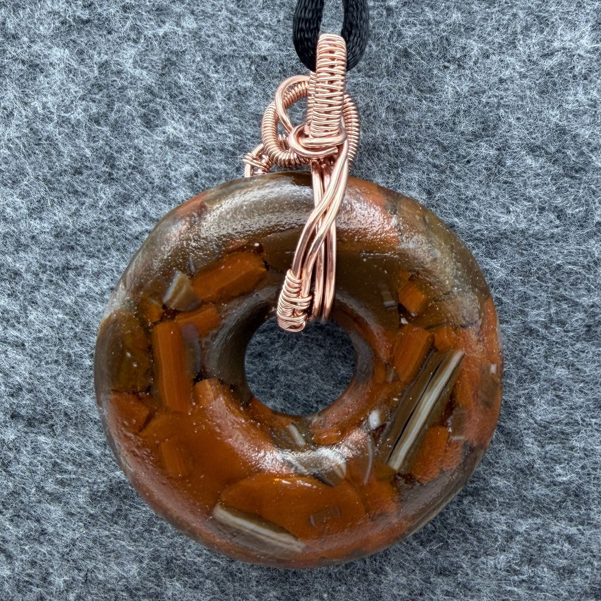 Back view of a fused glass donut shaped bead wrapped in copper wire held by a black cord against a gray felt background.