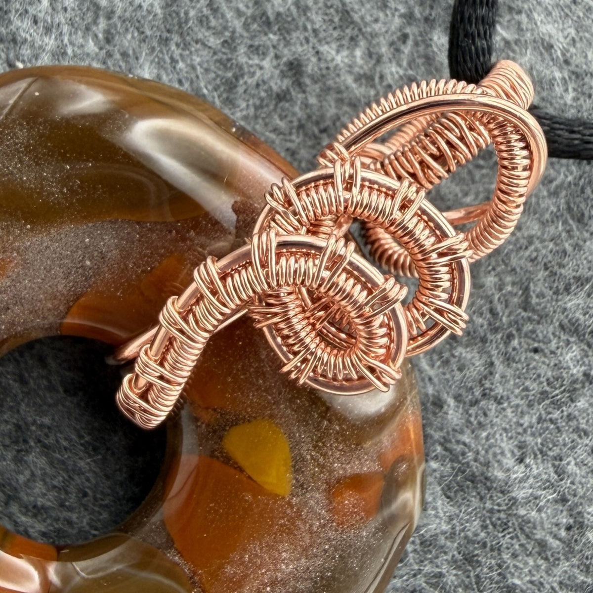 Detail view of a fused glass donut shaped bead wrapped in copper wire held by a black cord against a gray felt background.