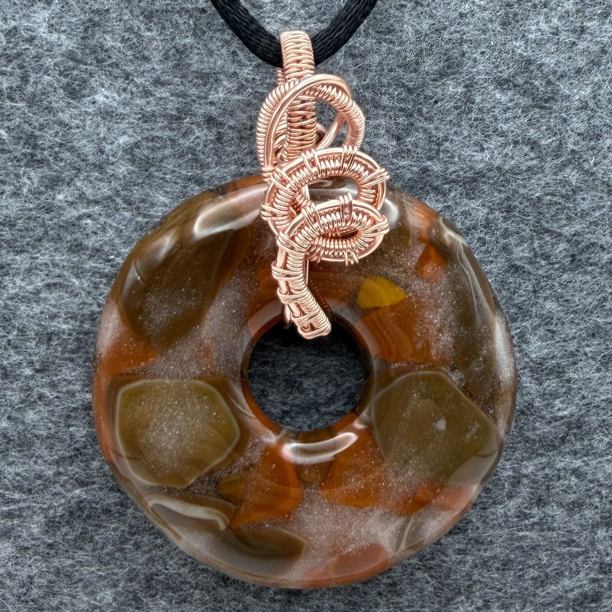 Front view of a fused glass donut shaped bead wrapped in copper wire held by a black cord against a gray felt background.