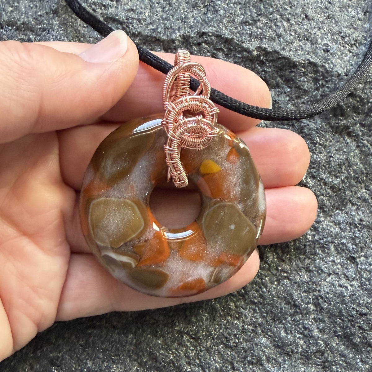 Front view of a fused glass donut shaped bead wrapped in copper wire suspended on a black cord held in the artist's hand.