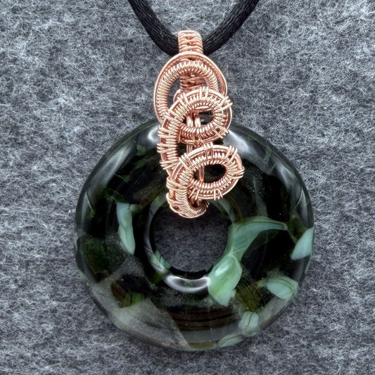 Front view of a fused glass donut bead wrapped in copper wire threaded through with a black satin cord against a gray felt background.