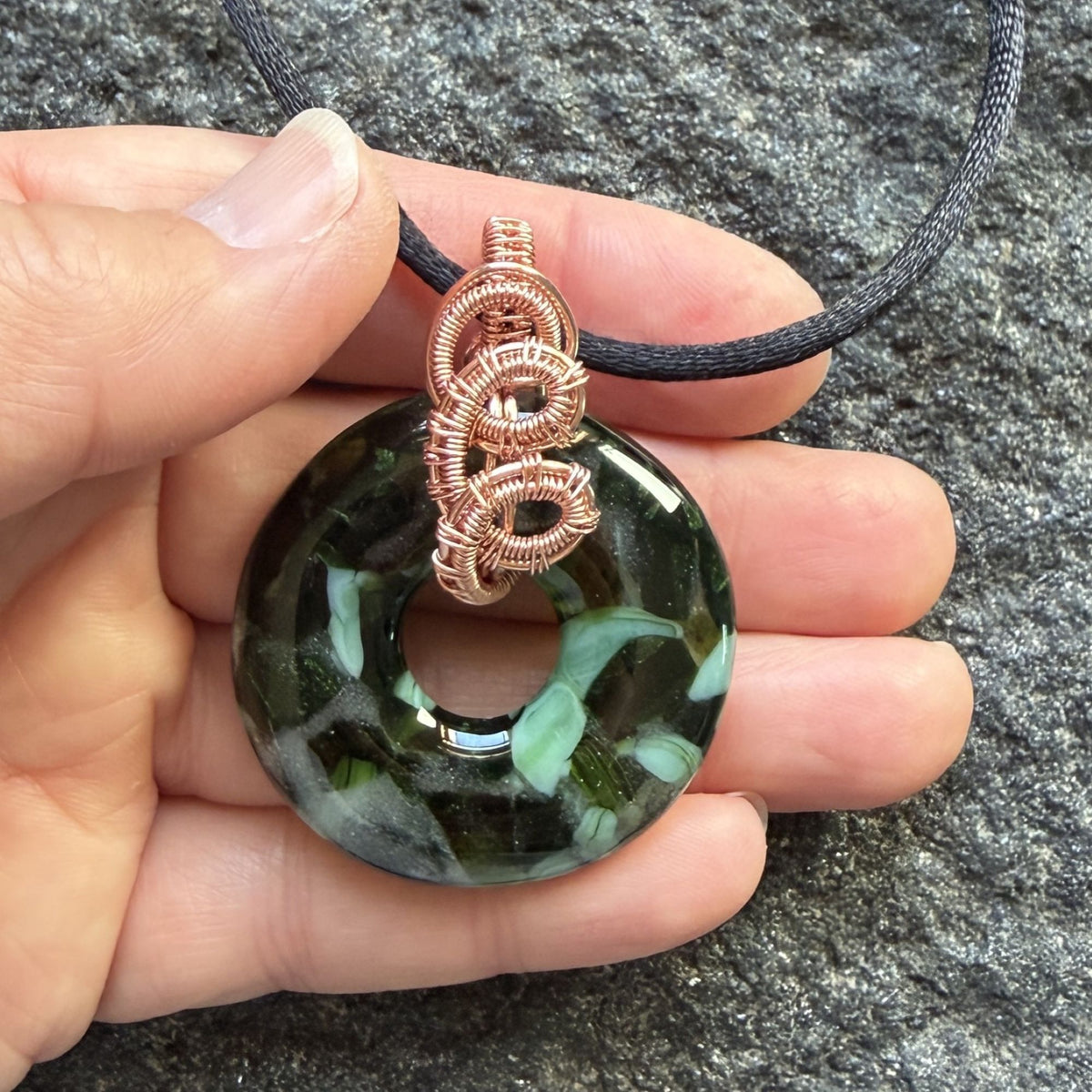 Front view of a fused glass donut bead wrapped in copper wire threaded through with a black satin cord held in the artist's hand.