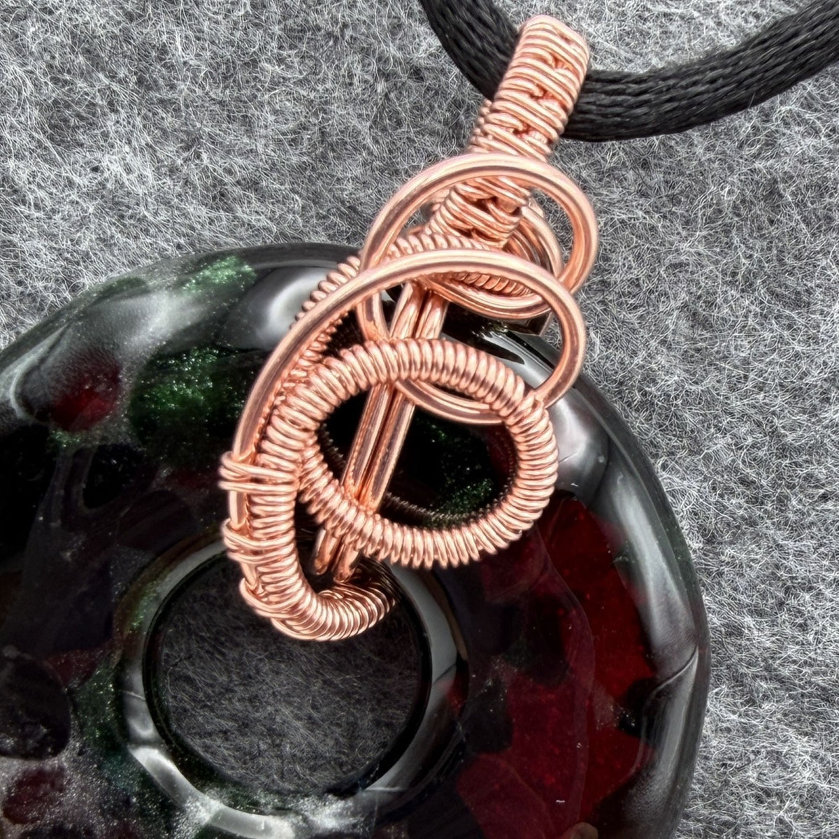 Detail view of a fused glass donut wrapped in copper wire hanging on a black satin cord against a gray felt background.