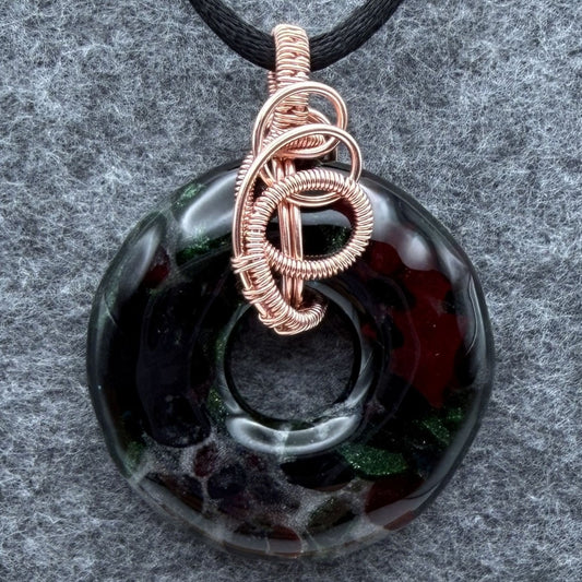 Front view of a fused glass donut wrapped in copper wire hanging on a black satin cord against a gray felt background.