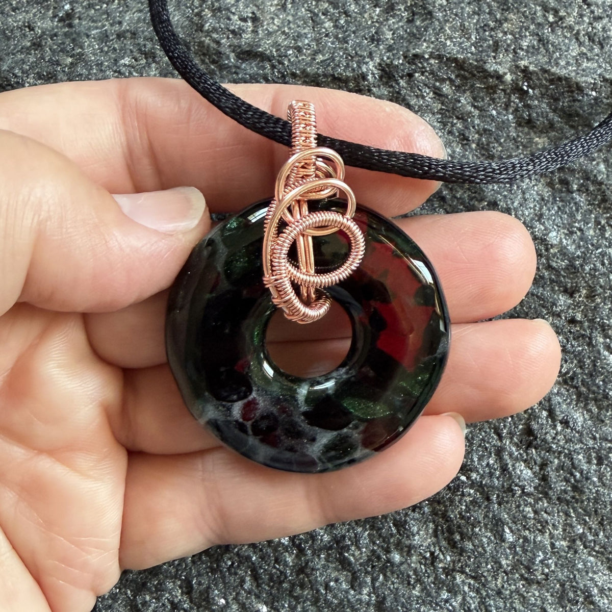 Front view of a fused glass donut wrapped in copper wire hanging on a black satin cord held in the artist's hand.